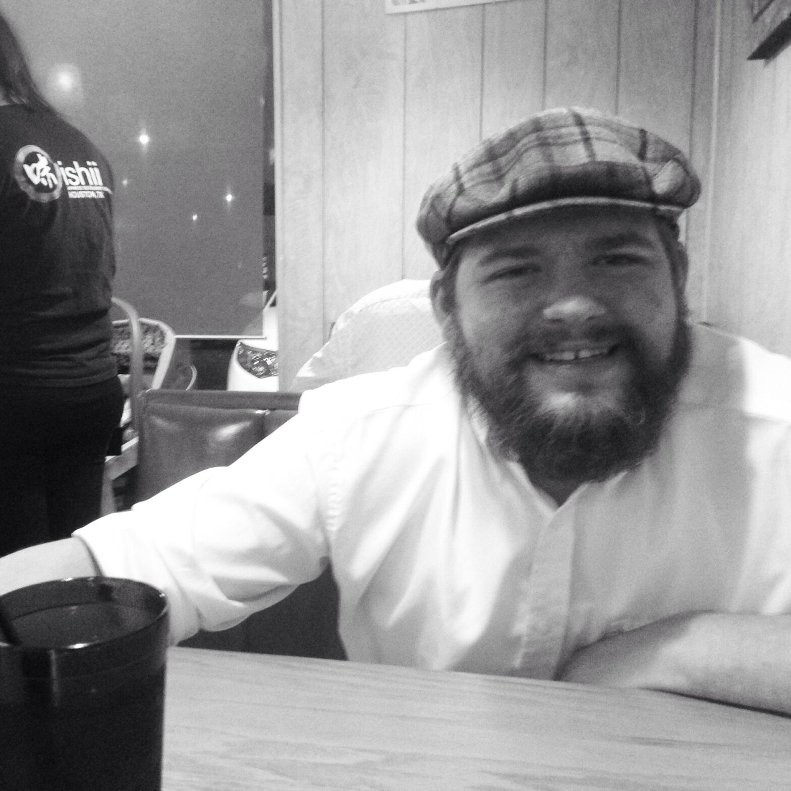 Black and white photo of author Mike Hilbig, bearded and wearing a flat cap. He's grinning.